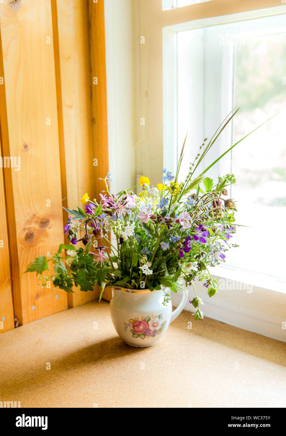 Vintage style flower bouquet made of wild flowers found in forest and meadow, standing in old cream jug on window sill, summers in grandma`s house con Stock Photo